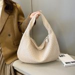 Sac à main tissé beige élégant, porté par une femme en manteau marron, posé sur un banc gris avec un livre blanc à côté. Mode féminine chic.